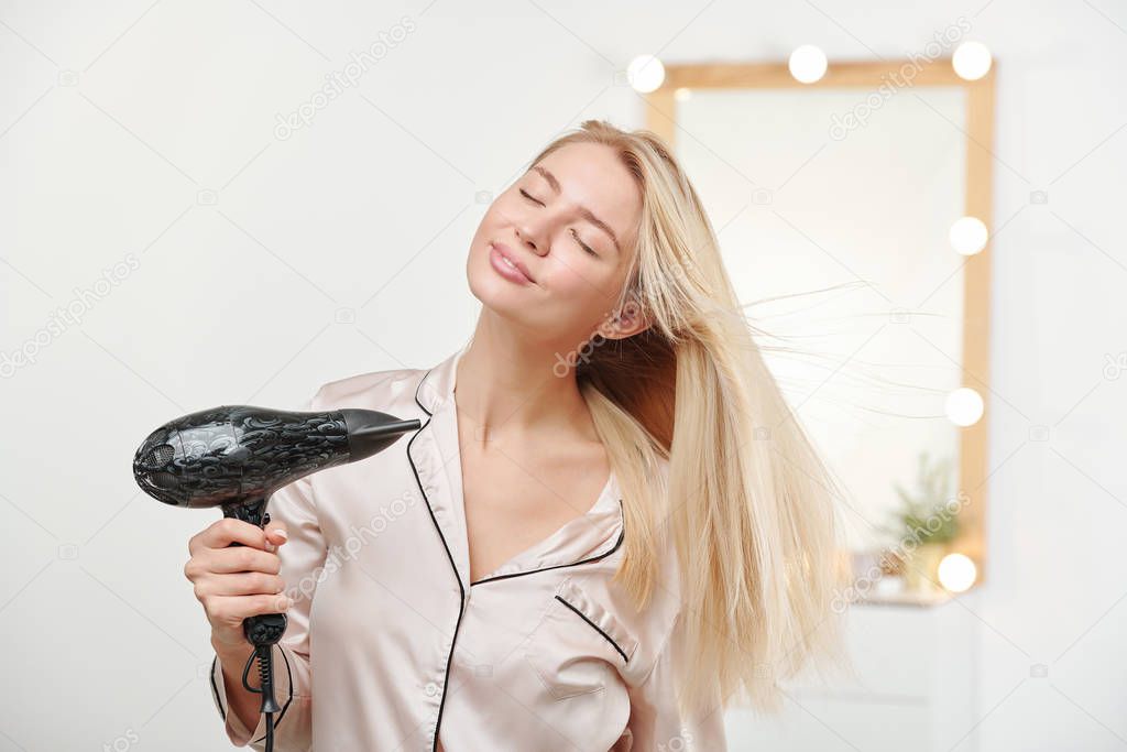 Young beautiful woman taking pleasure in warm air while using hairdryer after washing her long thick healthy blond hair