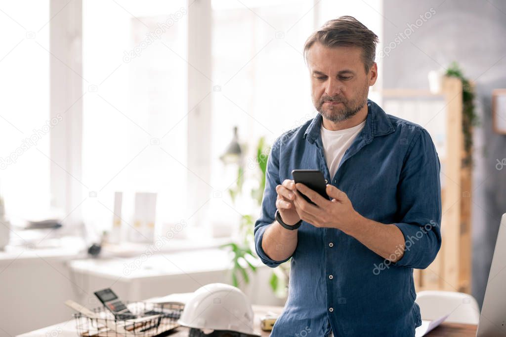 Serious engineer in casualwear scrolling through contacts in his smartphone while working in office