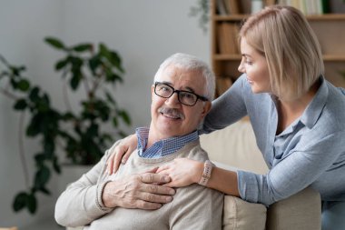 Restful senior man on couch holding hand of his young blonde daughter standing near by and embracing her father
