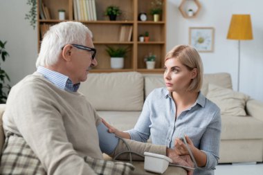 Young blonde careful daughter with tonometer looking at her senior father while measuring his blood pressure at home