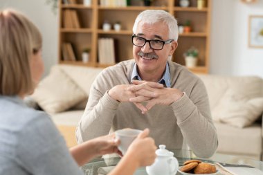 Senior retired man looking at his daughter with smile during conversation by cup of tea while sitting by table in living-room