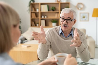 Retired man explaining something to his young daughter during discussion by cup of tea in living-room