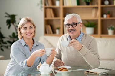 Smiling young woman and her senior father having tea by served table in front of camera in living-room
