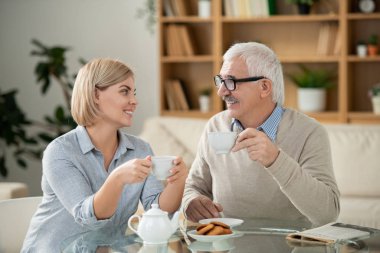 Cheerful young woman and her elderly father with cups of tea looking at each other while sitting by served table in front of camera