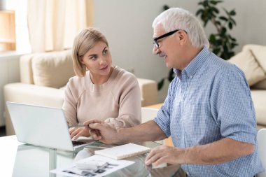 Aged businessman pointing at laptop display while looking at his young female colleague during discussion of online data