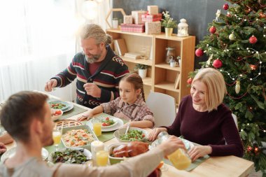Young man pouring orange juice for his happy mom sitting against decorated Christmas tree during family dinner at home