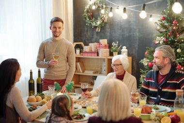 Cheerful young man with glass of wine toasting by served table in front of his family during Christmas dinner at home