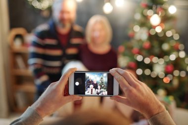Hands of man holding smartphone while taking photo of big relaxed family by festive dinner on Christmas eve