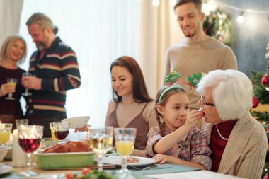 Adorable girl giving her great grandmother small tomato while both sitting by festive table on background of young and mature couples