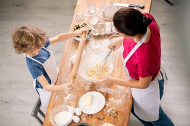 Little boy holding rolling-pin over wooden table while standing in front of his mother making dough for pastry clipart