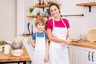 Adorable little boy and his mother in white aprons holding kitchenware while cooking together in the kitchen