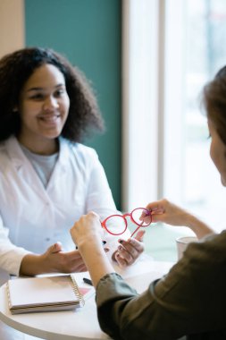 Happy young clinician sitting by table in front of patient and consulting her about new eyeglasses in clinics