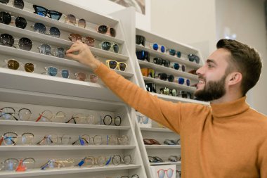 Happy young man taking pair of stylish eyeglasses while standing by display with eyewear assortment in optics shop