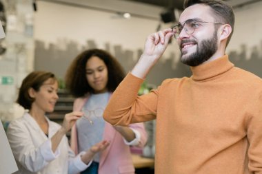 Happy guy trying on pair of eyeglasses on background of consultant helping young female client