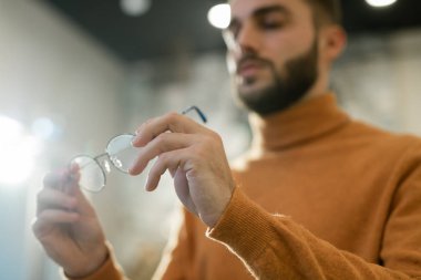 Hands of young man in yellow pullover holding pair of new eyeglasses while choosing eyewear in optics shop