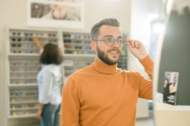 Young smiling man in pullover trying on new eyeglasses in front of mirror on background of female buyer by display with eyewear