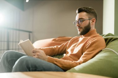 Young relaxed man in casualwear and eyeglasses sitting on comfortable sofa and reading book at leisure
