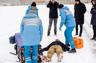 Brigade of young paramedics in winter uniform pushing stretcher towards unconscious man lying in snow