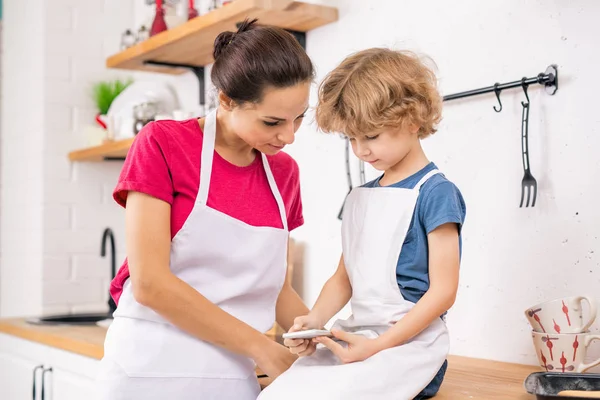 Adorable Blond Boy Apron Holding Smartphone While Showing His Mother — Stockfoto