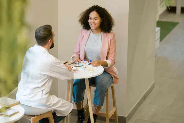 Pretty young client of optics shop or clinics sitting by table in front of clinician consulting her about choice of eyeglasses