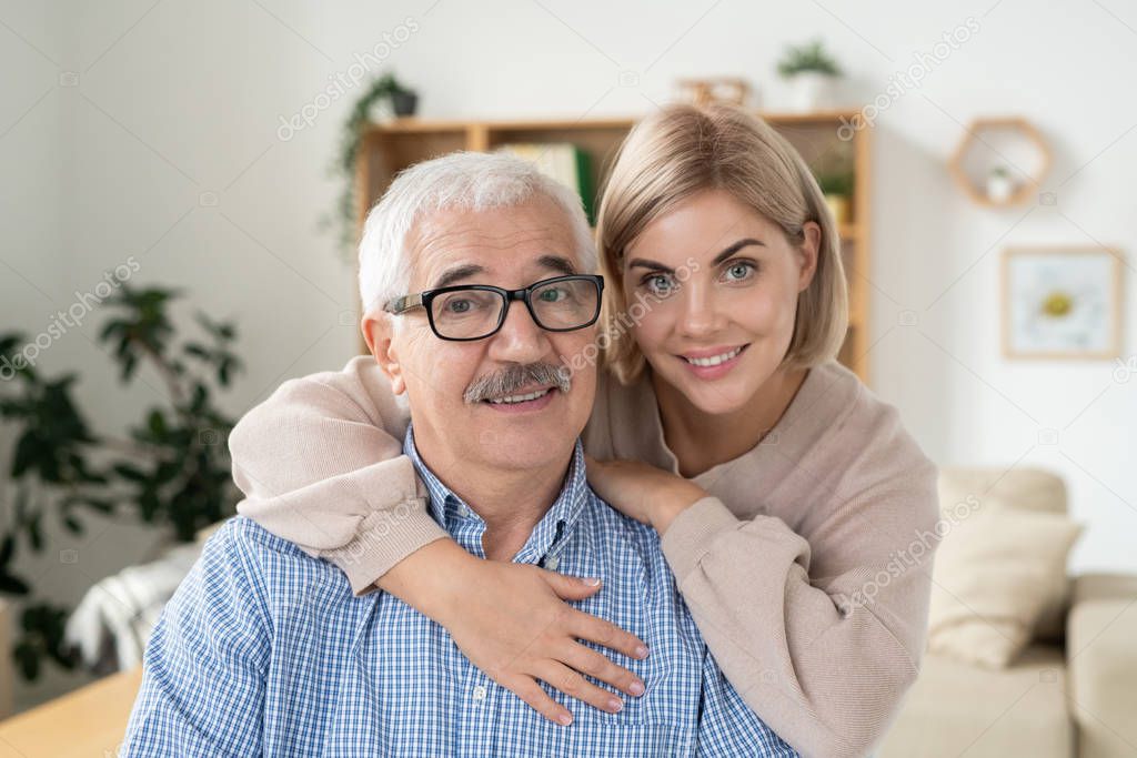 Young smiling blonde woman embracing her aged retired father in eyeglasses while both looking at camera at home