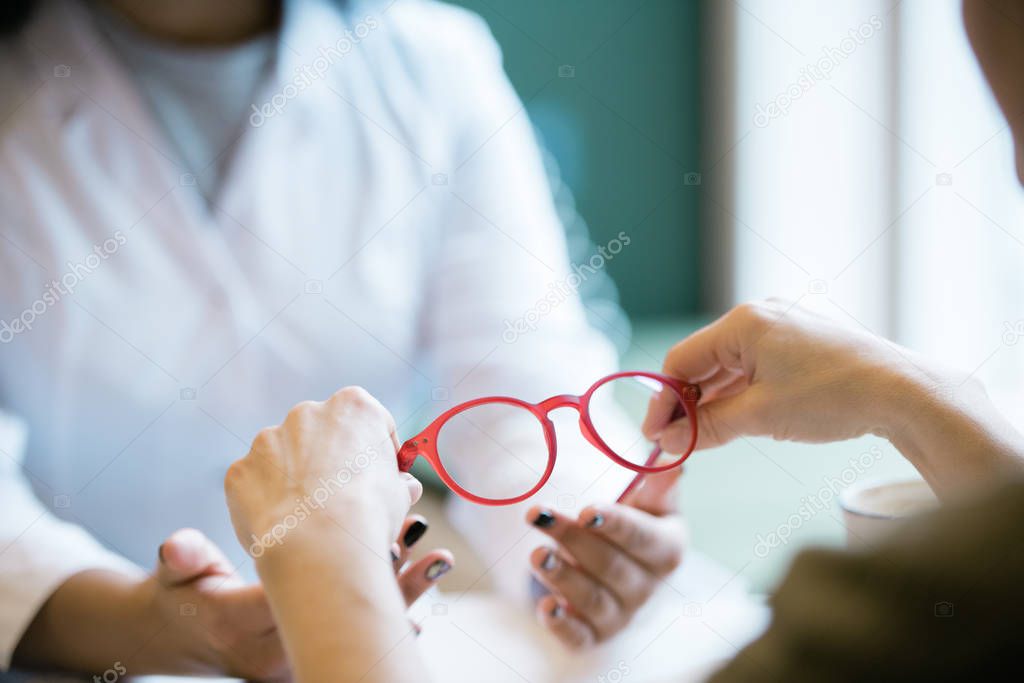 Hands of young female taking pair of new eyeglasses from those of optics clinician during consultation