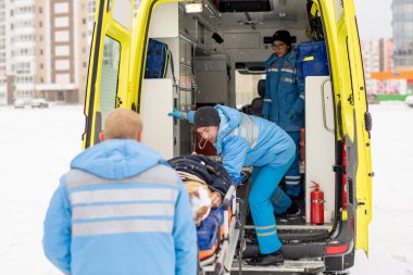 Paramedics in blue workwear getting stretcher with fixed unconscious man into ambulance car before going to hospital