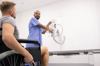 Young man with broken leg sitting on wheelchair and looking at radiologist standing by new medical equipment in clinics