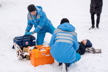 Two paramedics in uniform bending over sick unconscious man lying on snow while giving him first aid outdoors