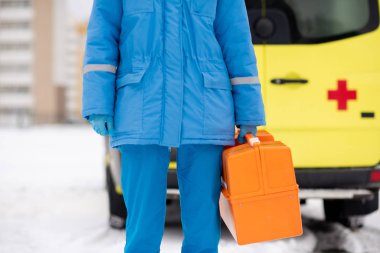 Mid-section of contemporary young paramedic in blue workwear and gloves holding first aid kit while standing in front of camera outdoors