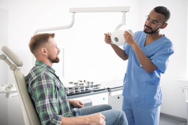 Young doctor in uniform holding medical equipment or lamp while standing in front of patient in armchair before examination