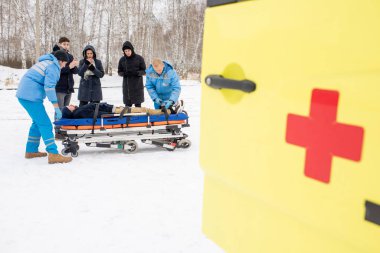 Two paramedics and three young people bending over stretcher with fixed unconscious man with ambulance car on foreground