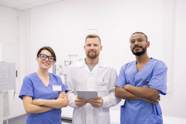 Group of three young successful intercultural clinicians or interns in uniform standing in front of camera in hospital