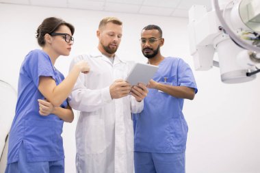 Group of three young clinicians in uniform consulting about peculiarities of new medical equipment while using tablet