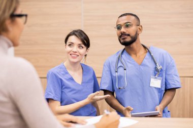 Pretty clinician in uniform smiling at patient while introducing her professional or colleague competent in important questions