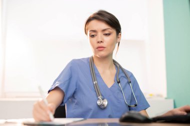 Young serious doctor or intern in blue uniform making working notes while sitting by workplace in front of computer