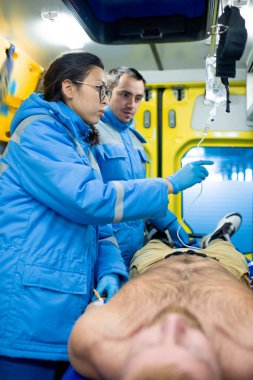 Two young paramedics standing by shirtless sick man inside ambulance car and preparing dropper for patient