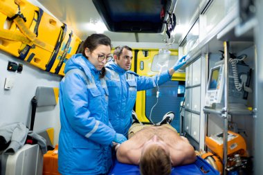 One of two paramedics examining sick unconscious man on stretcher while her colleague preparing dropper