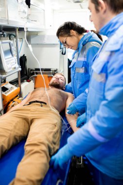 Young female paramedic in uniform giving first aid to sick shirtless man lying on stretcher inside ambulance car