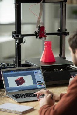 Genç işadamı dizüstü bilgisayarın önünde oturuyor ve basmadan önce yeni 3D modelin resmi üzerinde çalışıyor.