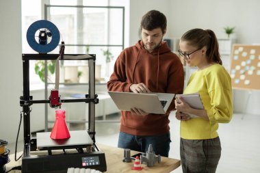 Yaratıcı proje için yeni online fikirler arayan iki genç yaratıcı tasarımcı 3D yazıcının önünde duruyor.
