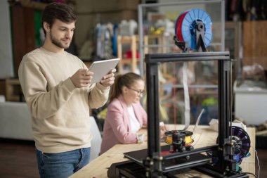 Touchpad 'li genç adam, meslektaşının arka planında 3D yazıcının önünde dikilirken çevrimiçi figüre bakıyor.