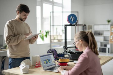 Dokunmatik bloknotu olan genç tasarımcı 3D yazıcının önünde dururken ve meslektaşı figürlerin taslağını çizerken internetten çalışıyor.