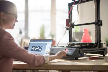 Yaratıcı kadın tasarımcı dizüstü bilgisayarın önünde otururken çalıştırmak için 3D yazıcının kontrol panelinde düğmeye basıyor