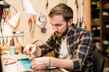 Sakallı tamirci, bozuk aletin üzerine eğiliyor ve tornavidayla küçük parçalarını ya da sökülmüş akıllı telefonun cıvatalarını tamir ediyor.