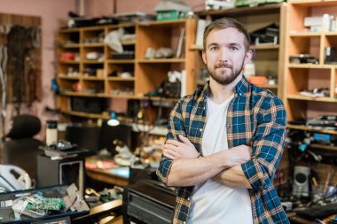 Young professional repairman of broken gadgets crossing arms by chest while standing in front of camera in his workshop clipart