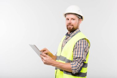 Beyaz kasketli, kareli gömlekli ve sarı yelekli, kameranın önünde tablet kullanan başarılı sakallı genç müteahhit.