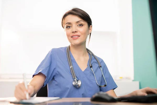 Pretty Young Smiling Doctor Blue Uniform Making Notes Workplace Front — 图库照片