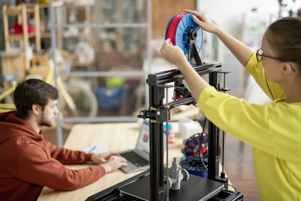 在3D打印机上 年轻的女职员正在更换带有红色长丝的卷轴 背景是同事关系网 — 图库照片