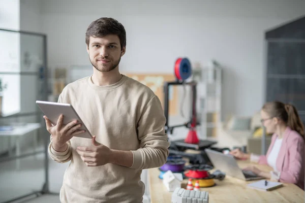 3Dプリンタで作業している同僚の背景にカメラの前に立つタッチパッドを持つ若いビジネスマン — ストック写真